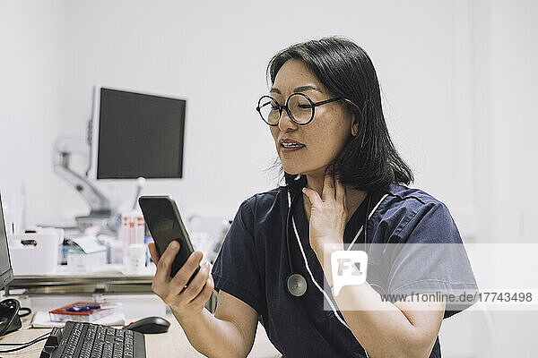 Eine HNO-Ärztin erklärt  während sie den Hals während einer Online-Konsultation über ein Smartphone in einer medizinischen Klinik berührt.