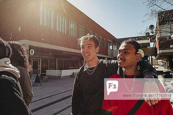 Young man with arm around male friend walking at street on sunny day