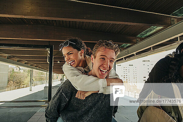 Happy young man piggybacking female friend at railroad station platform