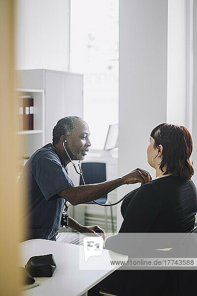Mature male doctor with stethoscope examining female patient sitting at desk in hospital