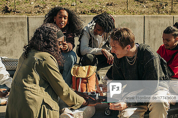 Lächelnder Mann trägt Nagellack auf die Hand einer Frau auf  während er mit Freunden im Park sitzt