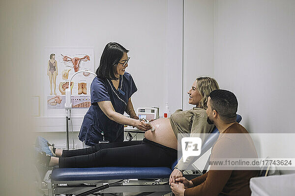 Lächelnde Ärztin bei der Ultraschalluntersuchung einer schwangeren Frau auf dem Untersuchungstisch eines Mannes im Krankenhaus