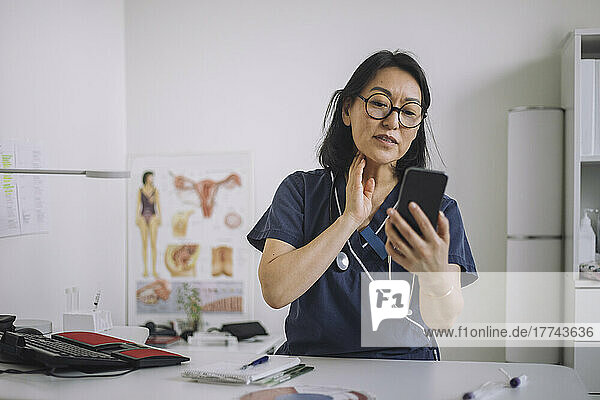 Weibliche HNO-Ärztin bei der Online-Konsultation über das Mobiltelefon  während sie am Schreibtisch in einer medizinischen Klinik sitzt