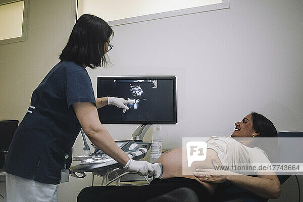 Weiblicher Gynäkologe  der auf den Bildschirm zeigt  während er eine schwangere Frau in einer medizinischen Klinik mit Ultraschall untersucht