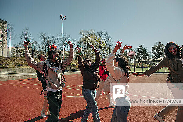 Unbeschwerte multirassische Freunde mit erhobenen Armen tanzen zusammen auf dem Sportplatz an einem sonnigen Tag