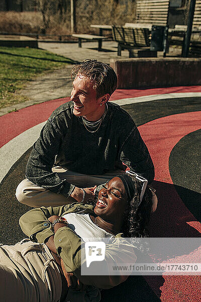 Lächelnde Frau  die auf dem Schoß eines männlichen Freundes liegt und sich auf einem Spielplatz entspannt