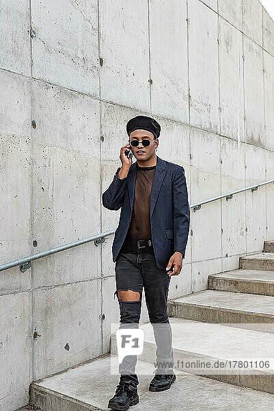Young Latin gay man wearing a fashionable hat and sunglasses  walking down the stairway while talking on his smartphone  before a white background. LGTB. Copy space