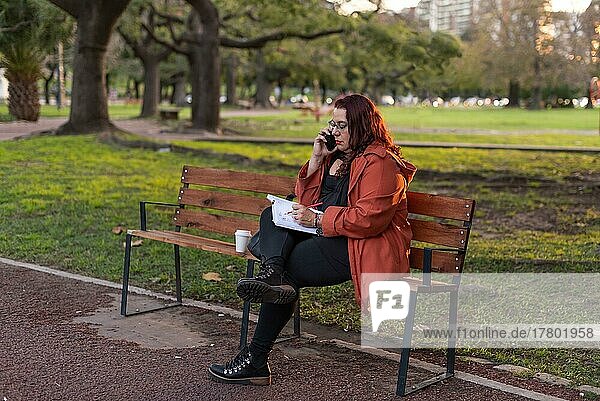 Eine leitende Frau in Übergröße arbeitet im Freien. Sie schreibt in ihr Notizbuch und führt ein Telefongespräch  während sie auf einer Bank in einem öffentlichen Park sitzt