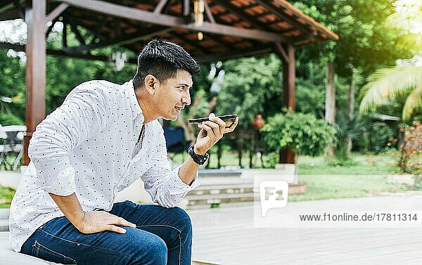 Hübscher Mann sitzt draußen und sendet eine Sprachnachricht mit seinem Handy  Teenager sitzt und telefoniert draußen  Attraktiver Mann sitzt und sendet eine Sprachnachricht mit seinem Handy und gestikuliert