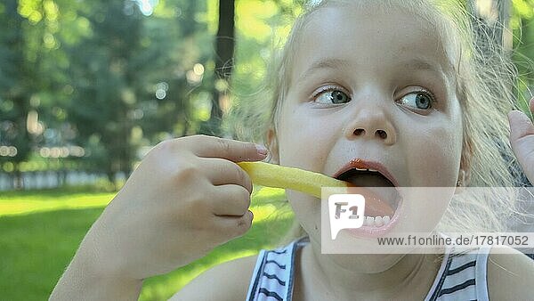 Kleines Mädchen essen Pommes frites. Close-up von blonden Mädchen nimmt Kartoffelchips mit ihren Händen und probiert sie sitzen in Straßencafé auf dem Park. Odessa  Ukraine  Europa