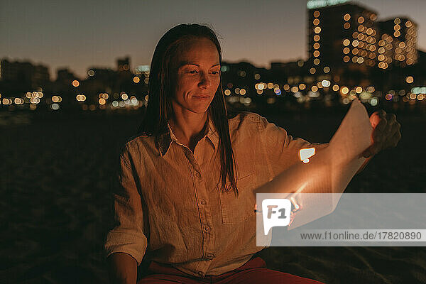 Woman with burning paper sitting on beach at night