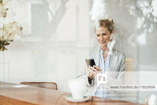 Smiling businesswoman looking at smart phone in cafe seen through glass