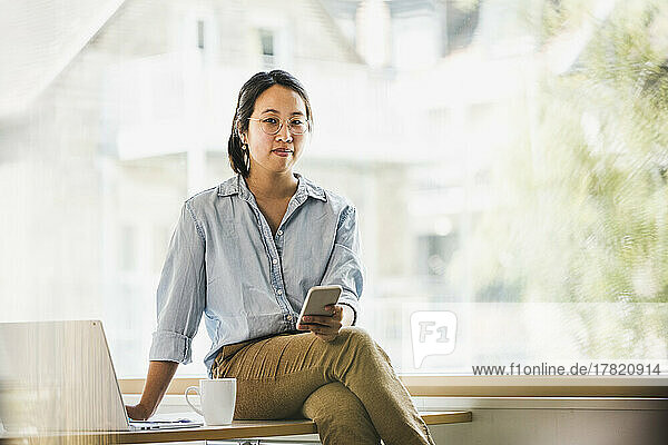 Smiling businesswoman with smart phone sitting on desk at work place