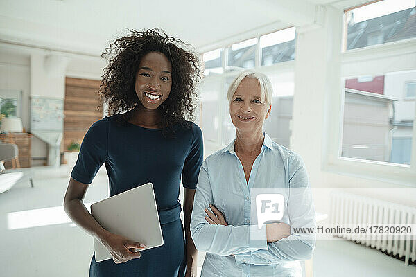 Smiling businesswoman holding laptop standing with colleague in office