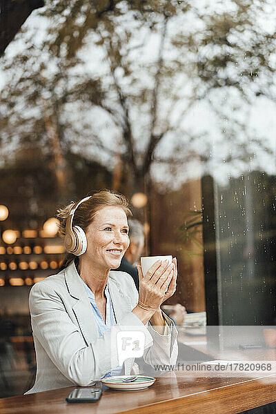 Smiling businesswoman holding coffee cup listening music in cafe seen through glass