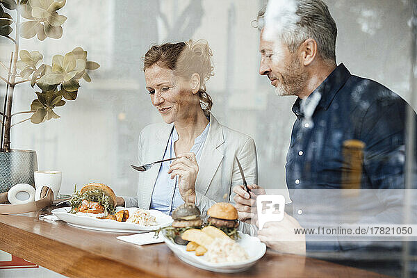 Smiling businesswoman with businessman having lunch in cafe seen through glass