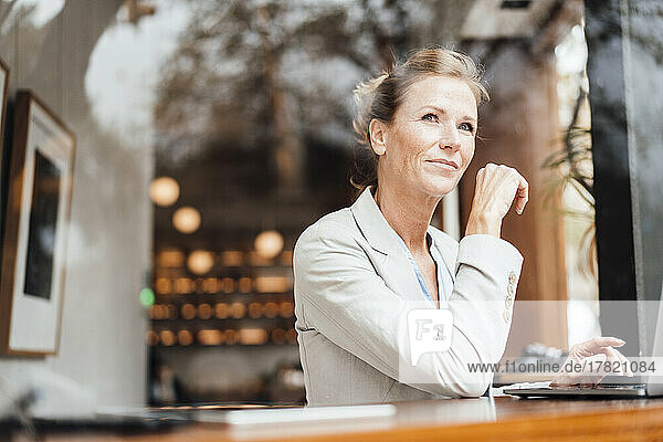 Smiling businesswoman sitting with laptop in cafe seen through glass