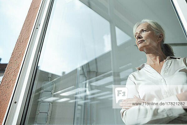 Thoughtful businesswoman standing with arms crossed seen through glass