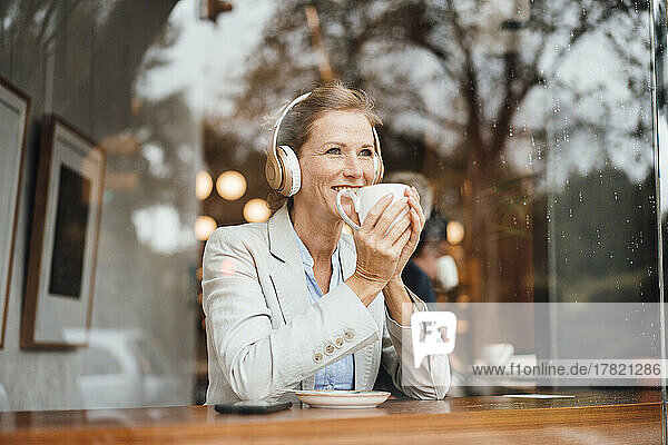 Smiling businesswoman holding coffee cup listening music through wireless headphones in cafe