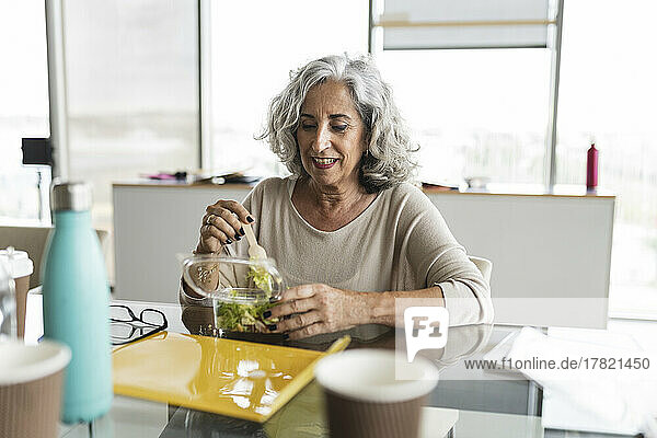 Happy businesswoman with gray hair eating salad at desk in office