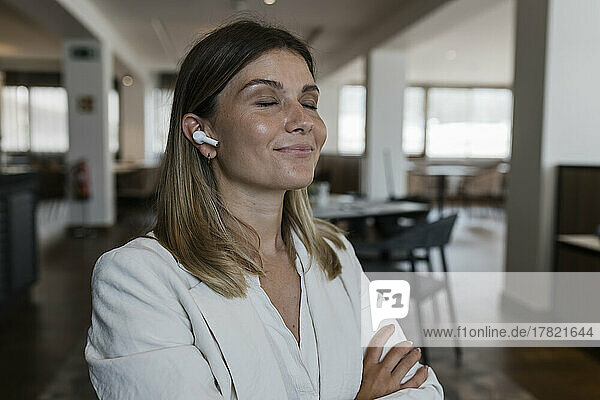 Smiling businesswoman with eyes closed listening music through wireless in-ear headphones at restaurant
