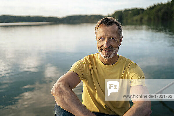 Happy mature man sitting in front of lake on sunny day