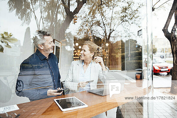 Smiling businesswoman holding credit card talking with man in cafe seen through window