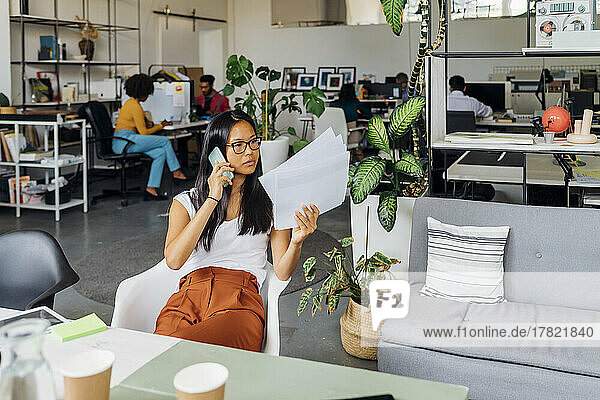 Junge Geschäftsfrau mit Papierdokumenten  die im Büro über ihr Mobiltelefon spricht