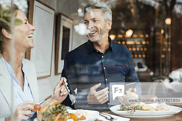 Happy businesswoman with businessman holding mobile phone having lunch in cafe seen through glass