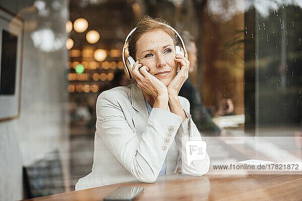 Businesswoman with head in hands listening music at cafe seen through glass