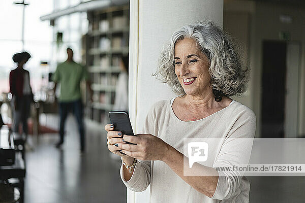 Smiling businesswoman using smart phone by column at office