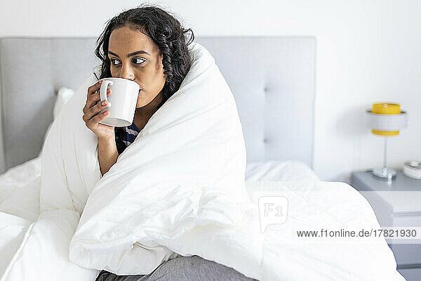 Woman drinking coffee looking away in bedroom