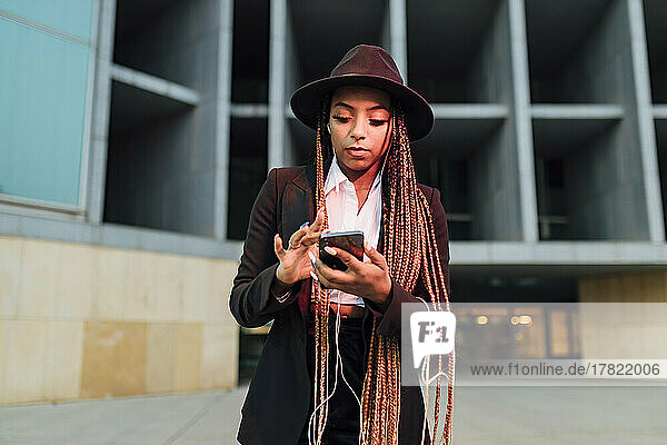 Businesswoman with smart phone listening music through in-ear headphones outside office building