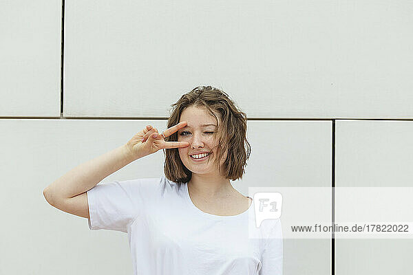 Lächelndes Teenager-Mädchen zwinkert mit den Augen und gestikuliert ein Friedenszeichen  das vor der Wand steht