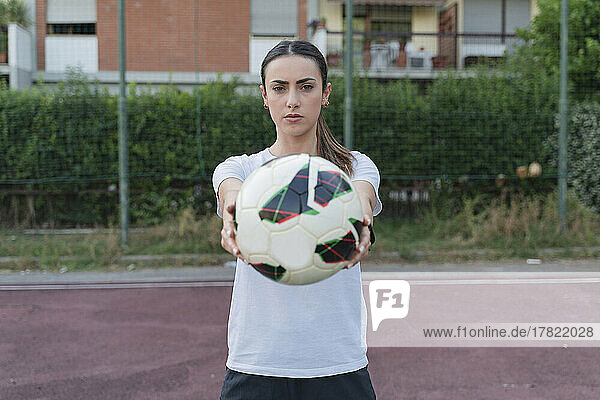 Confident young woman showing ball standing in soccer court