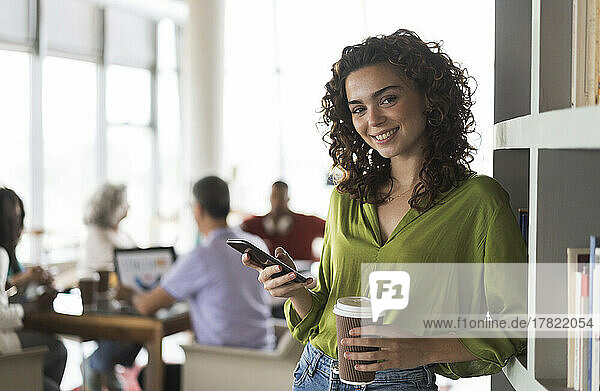 Smiling businesswoman with smart phone and disposable cup leaning on rack at office