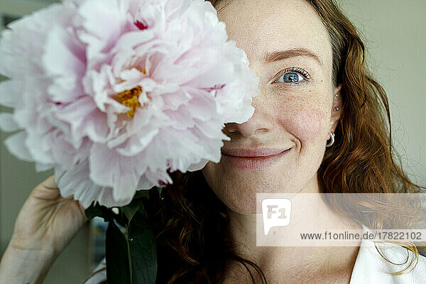 Smiling woman covering face with pink peony flower at home