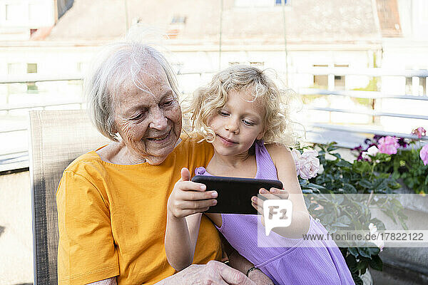 Lächelnde Enkelin zeigt der Großmutter auf dem Balkon ihr Mobiltelefon