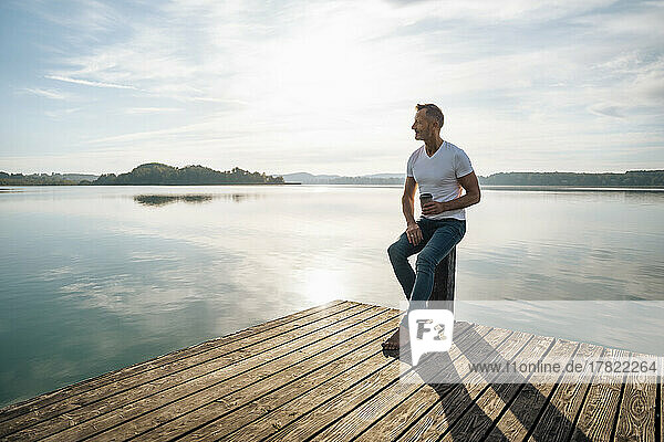 Mature man having coffee sitting on pier by lake