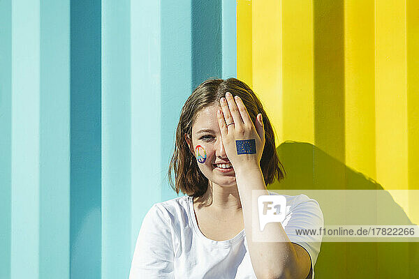 Lächelndes Teenager-Mädchen mit Friedenssymbolfarbe im Gesicht  das das Auge vor einer blauen und gelben Wand bedeckt