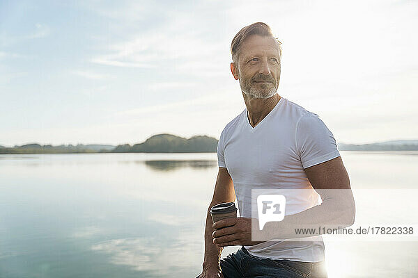 Thoughtful mature man sitting with coffee cup near lake on sunny day