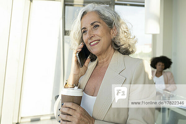 Ältere Geschäftsfrau hält Einwegbecher in der Hand und spricht im Büro mit dem Smartphone