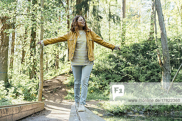 Smiling woman with arms outstretched walking on wooden plank in forest