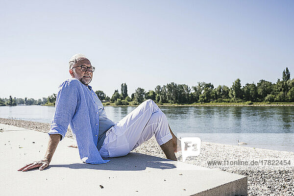 Älterer Mann mit gekreuzten Beinen am Flussufer an einem sonnigen Tag
