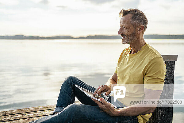 Happy mature man sitting with tablet PC on jetty at lake