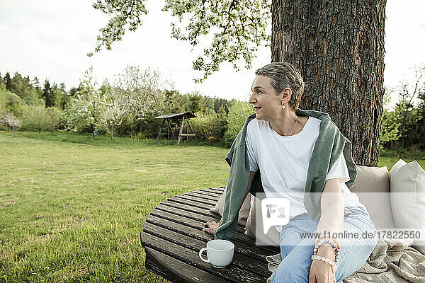 Lächelnde Frau mit Kaffeetasse sitzt auf Bank vor Baumstamm im Garten