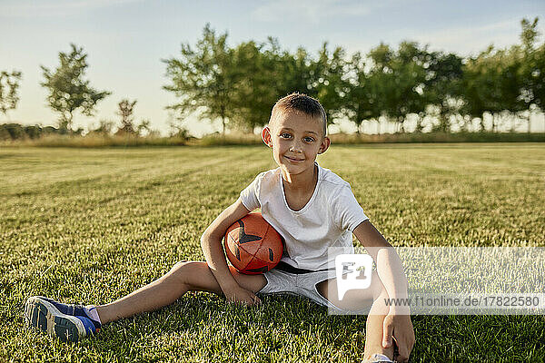 Smiling boy with rugby ball sitting at sports field on sunny day
