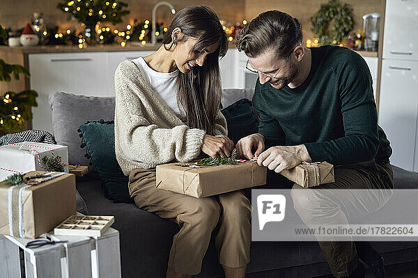 Smiling woman with man wrapping Christmas gift sitting on sofa at home