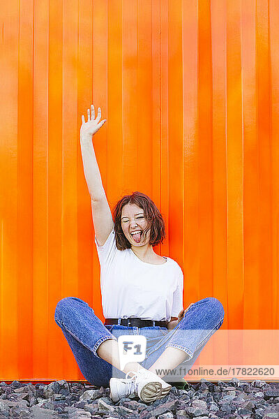 Fröhliches Teenager-Mädchen mit erhobener Hand und herausgestreckter Zunge sitzt vor einem orangefarbenen Frachtcontainer