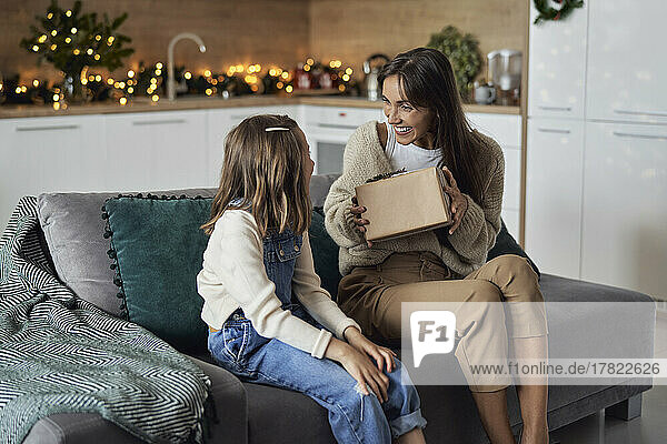 Smiling woman showing gift to daughter sitting on sofa at home